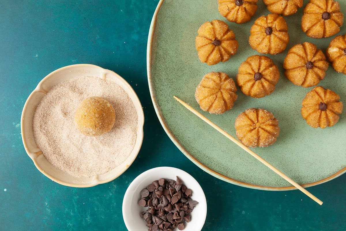 A plate of pumpkin-shaped cookies sits beside a bowl of sugar, a rolling stick, and a small dish of chocolate chips on a green surface. One cookie is being rolled in sugar.