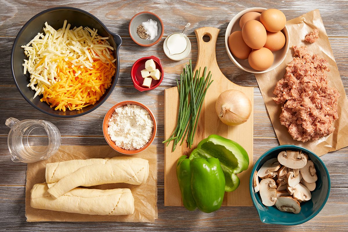 Overhead view of various omelet ingredients on a wooden surface, including shredded cheese, eggs, ground meat, mushrooms, green pepper, onion, flour, butter, chives, spices, water, and two rolled tortillas.