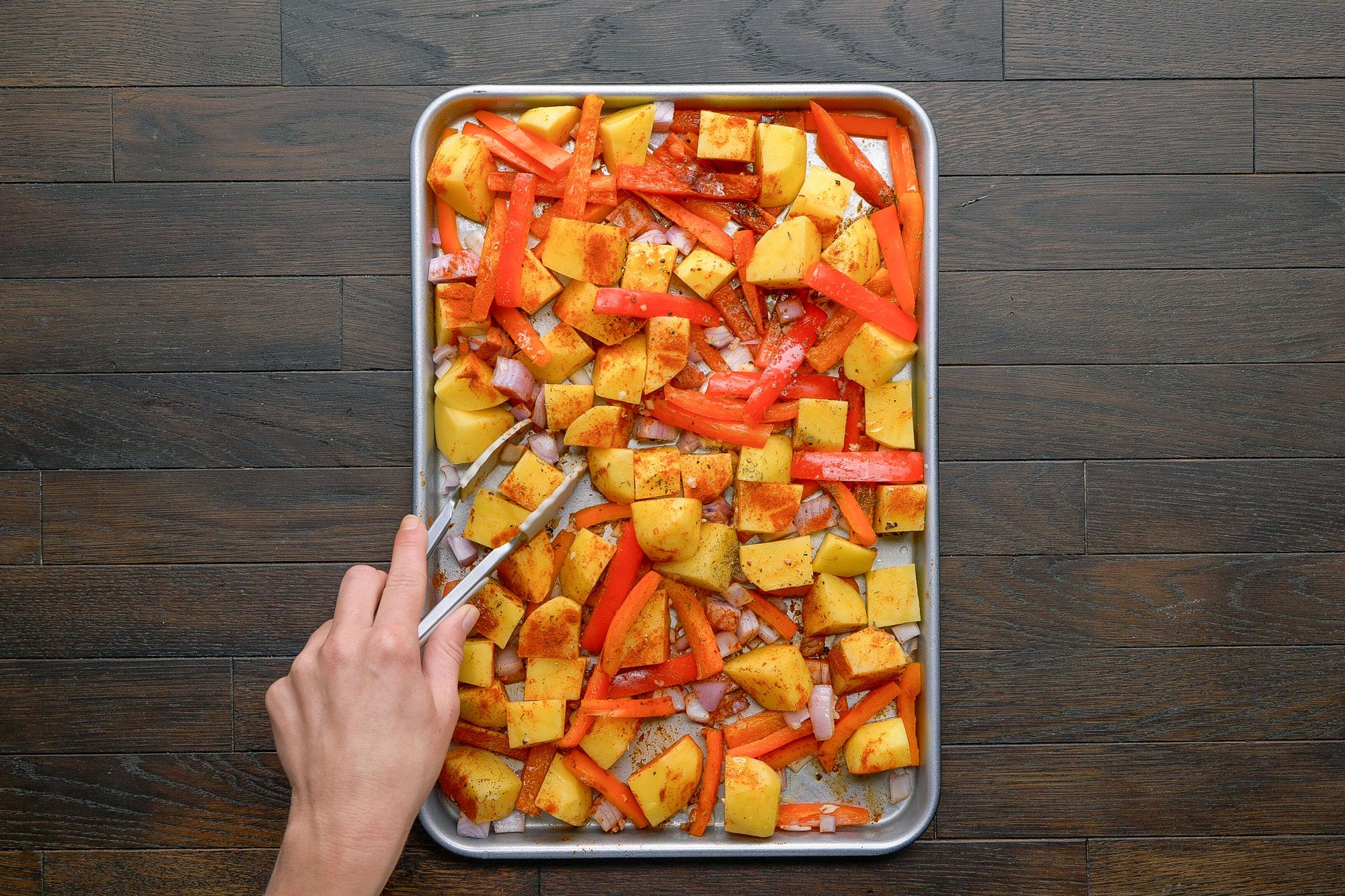overhead shot of a hand uses tongs to mix chopped potatoes, carrots, and onions seasoned with spices on a baking sheet, set on a dark wooden surface