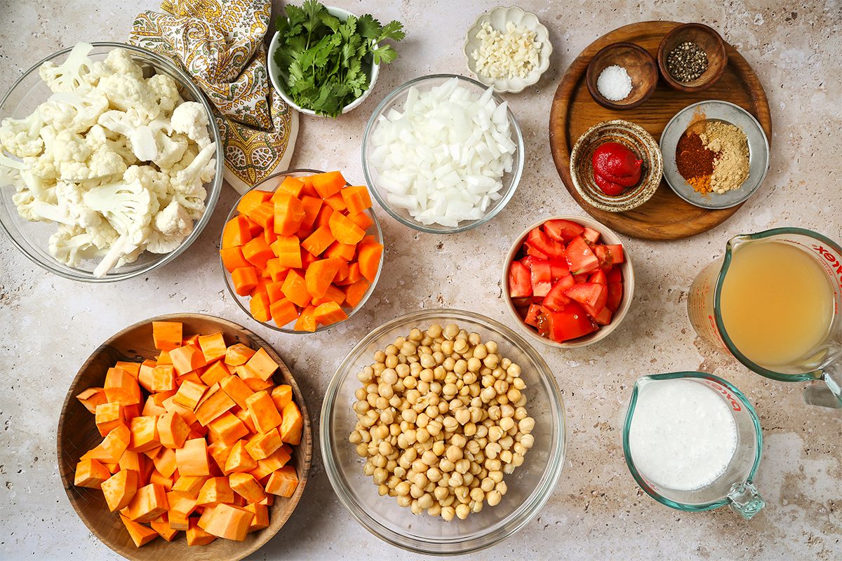 Various ingredients in bowls on a countertop, including chopped cauliflower, carrots, sweet potatoes, onions, tomatoes, chickpeas, garlic, fresh cilantro, spices, tomato paste, salt, pepper, broth, and coconut milk.