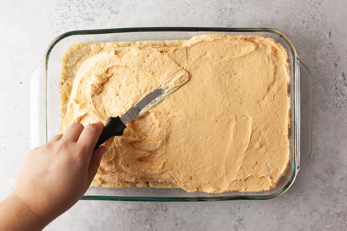 A hand uses an offset spatula to spread creamy frosting over a rectangular cake in a glass baking dish on a light countertop.