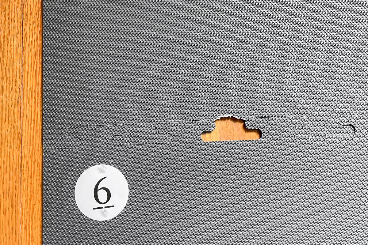 A textured gray panel with a puzzle-like connection slot and a piece missing, revealing a wooden surface beneath. A white circle with the number 6 is in the lower left corner of the panel.