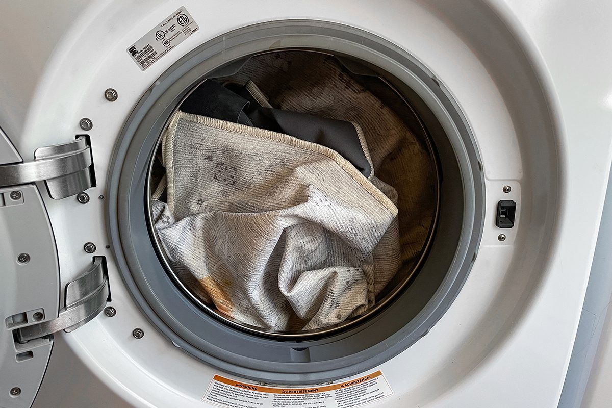 Front-loading washing machine with its door open, showing dirty or stained light-colored fabric inside the drum, ready to be washed.