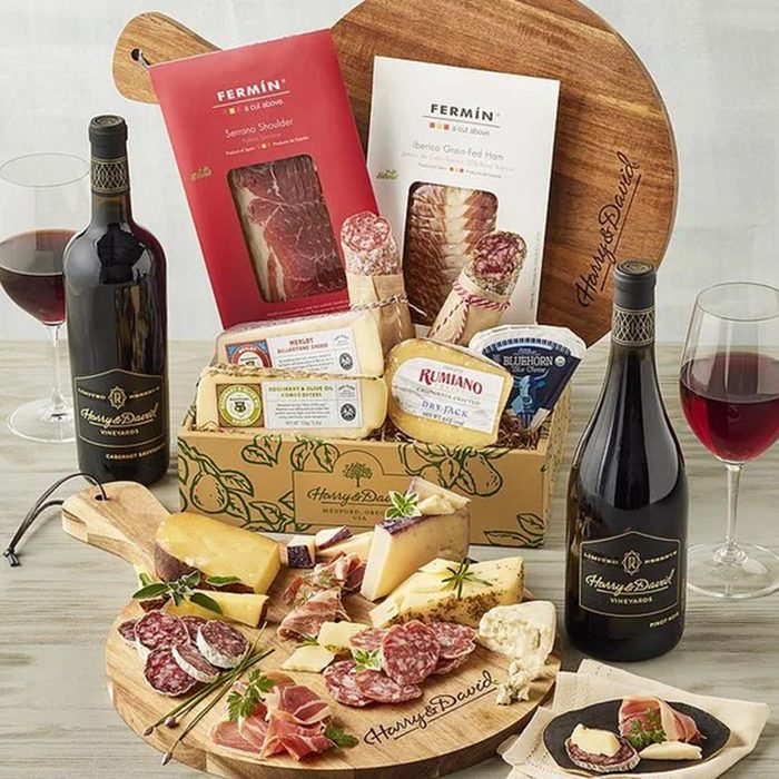 A wooden board displays assorted cheeses, cured meats, and crackers, surrounded by two bottles and two glasses of red wine, packaged meats, and more cheese, all arranged on a light-colored table.