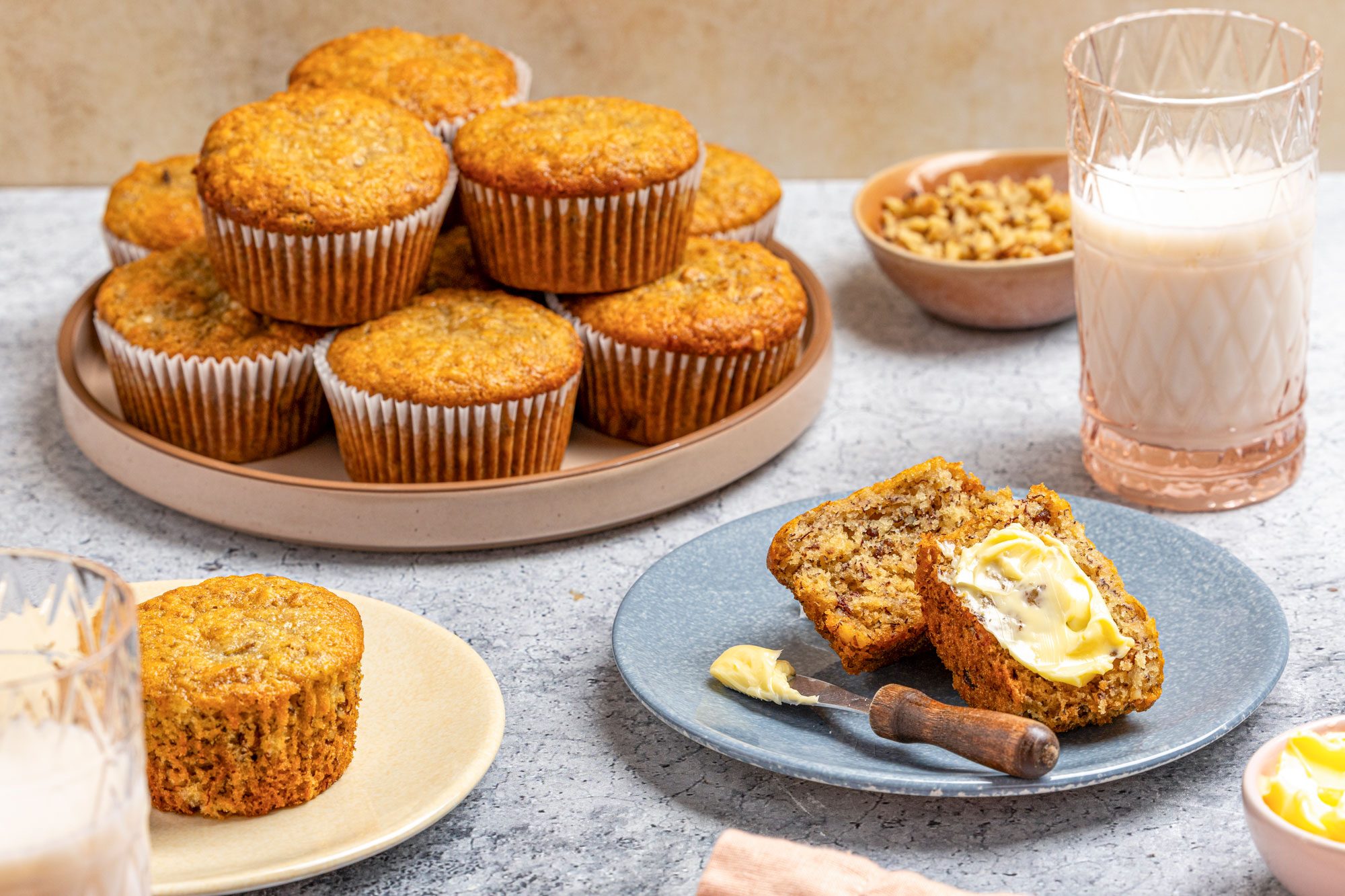 Table view shot of vegan banana muffins; A blue plate holds a halved muffin spread with butter and a knife, with a glass of milk and a bowl of chopped nuts nearby; More muffins in paper liners are stacked on a round tray, with an additional muffin on a beige plate; A small bowl of butter and a peach napkin rest on the gray surface