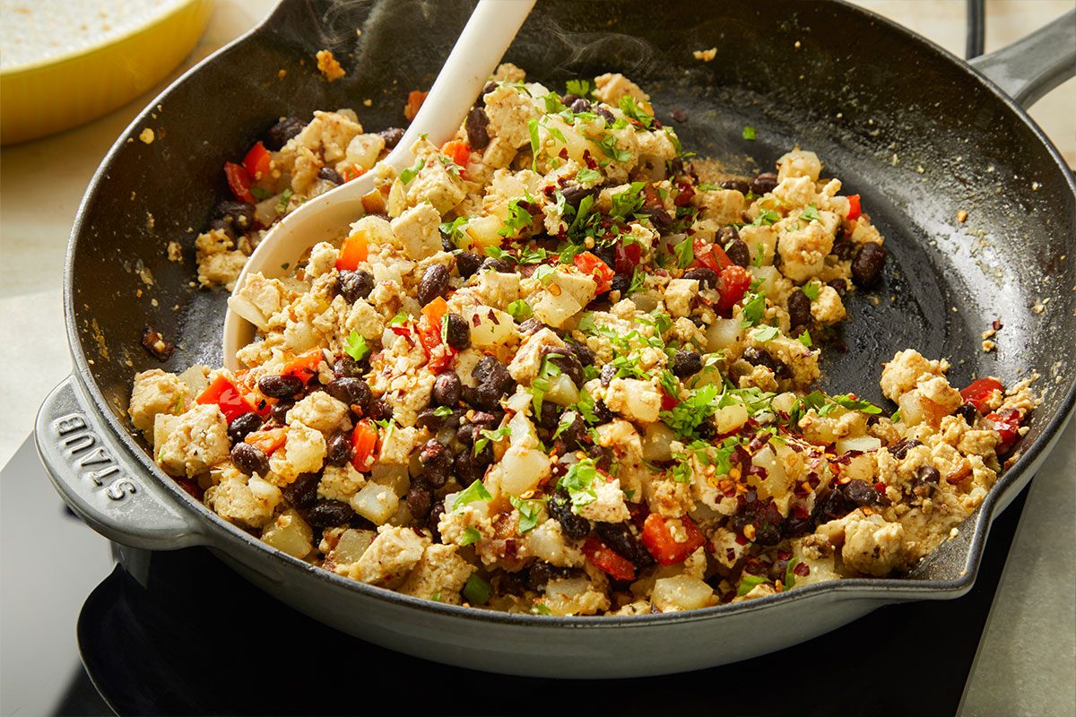 A skillet filled with a sautéed mixture of chopped vegetables, beans, and possibly tofu or potatoes, being stirred with a white spoon on a stovetop.