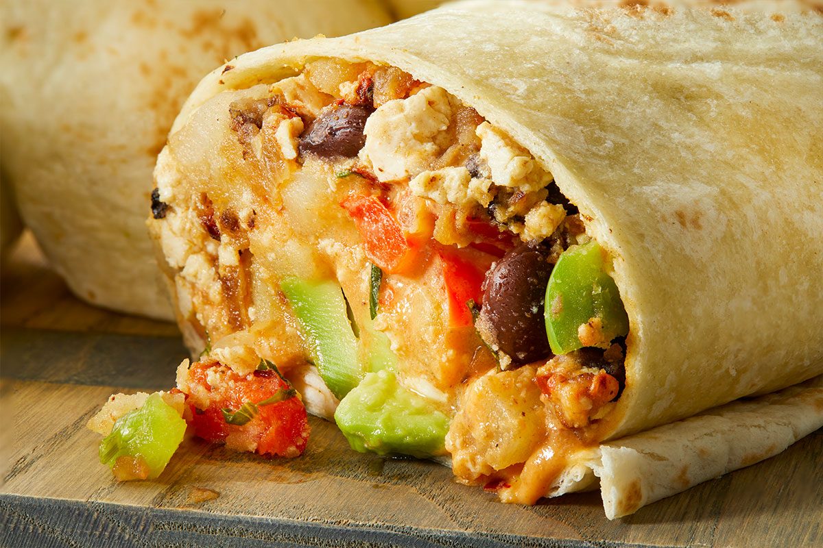 A close-up of a burrito cut open, showing a filling of beans, rice, diced green and red peppers, cheese, and pieces of chicken, wrapped in a flour tortilla on a wooden surface.