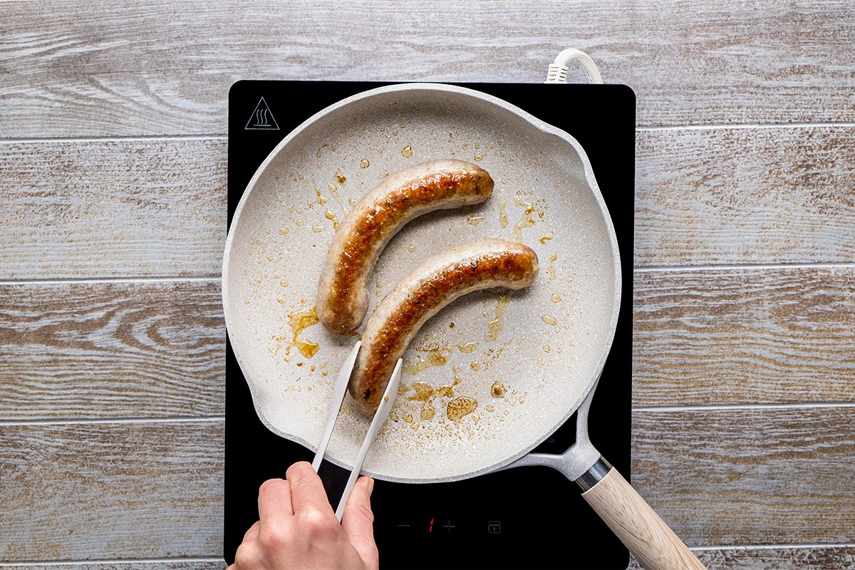 A hand uses tongs to cook two browned sausages in a white frying pan on an electric stovetop, with oil splatters visible in the pan. The stovetop sits on a light wood surface.