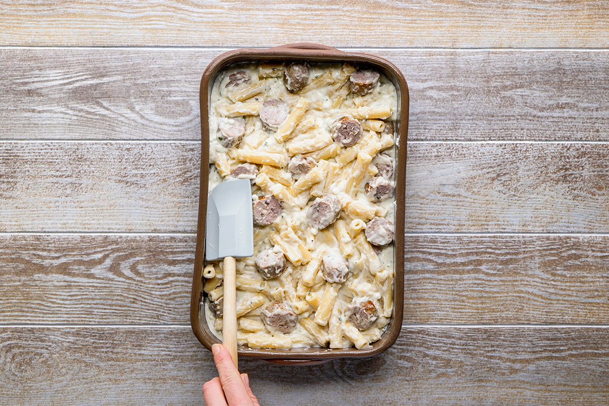 A hand holding a spatula is spreading a creamy baked pasta dish with sausage slices in a rectangular baking dish on a wooden surface.
