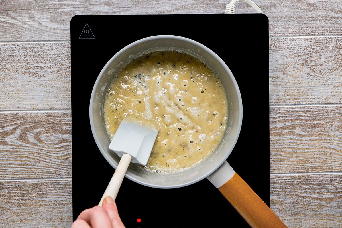 A hand stirs a bubbling, light brown mixture in a saucepan with a white spatula on an electric stovetop, atop a light wood surface.