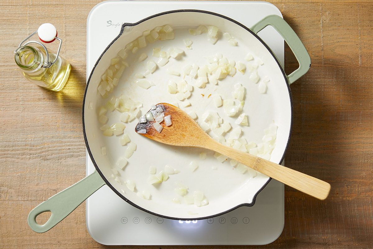 A white pan with chopped onions being sautéed, stirred with a wooden spoon, sits on a white stovetop. A small bottle of oil is on the wooden counter nearby.