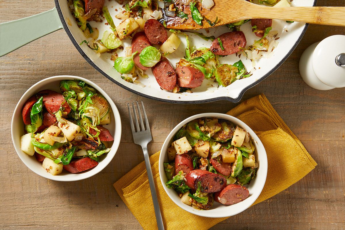 Two bowls of sautéed sausage, potatoes, and Brussels sprouts, with a pan of the same dish nearby. A fork rests on a yellow napkin beside the bowls, all set on a wooden table.