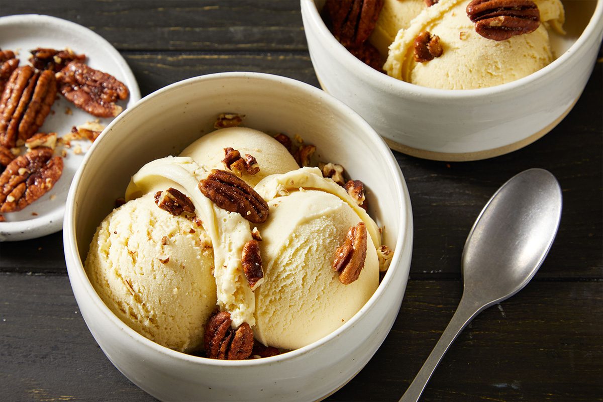 3/4th closeup shot of A bowl of creamy Bourbon ice cream topped with candied pecans sits on a dark wooden surface, with a spoon nearby and an extra dish of pecans in the background