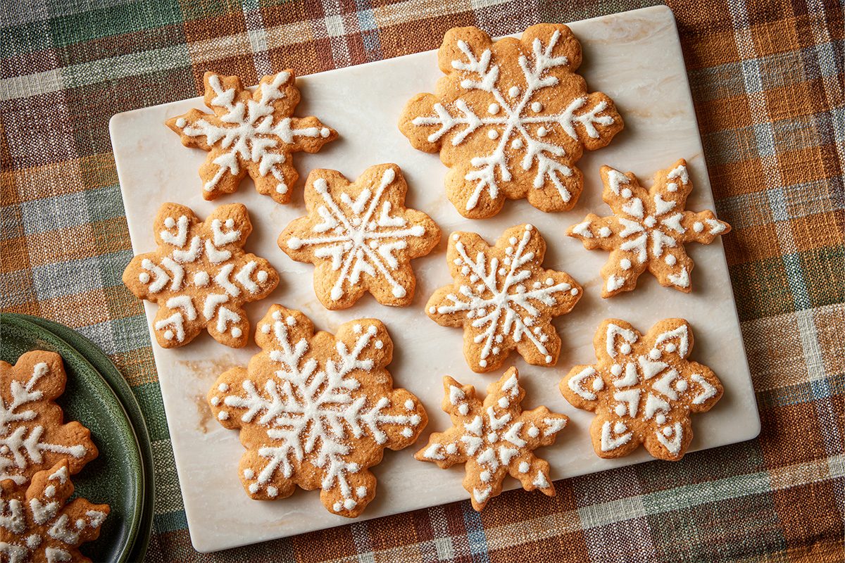 Overhead shot of a marble tray holding ten butterscotch gingerbread cookies shaped like snowflakes with white icing, set on a warm plaid tablecloth;