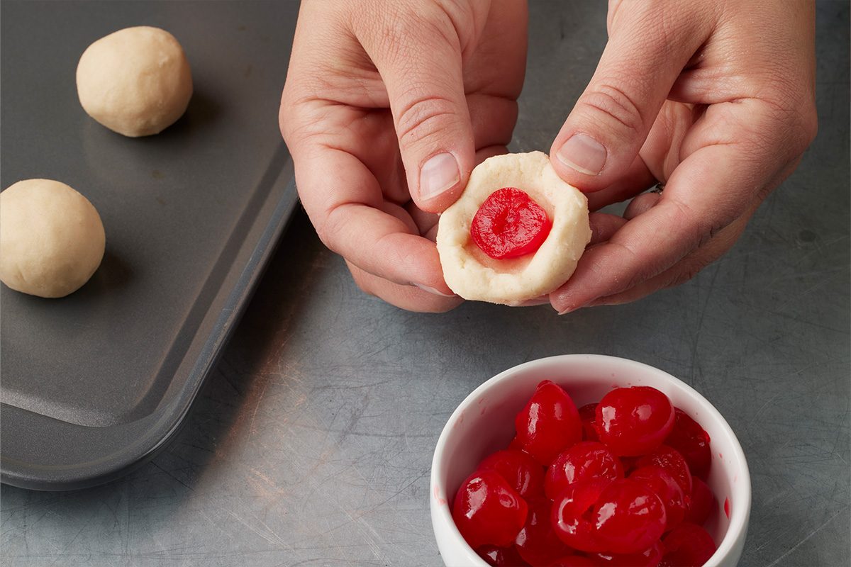 Closeup shot of hands shaping cookie dough around a red cherry, with a bowl of cherries and a baking sheet of dough balls in the background;