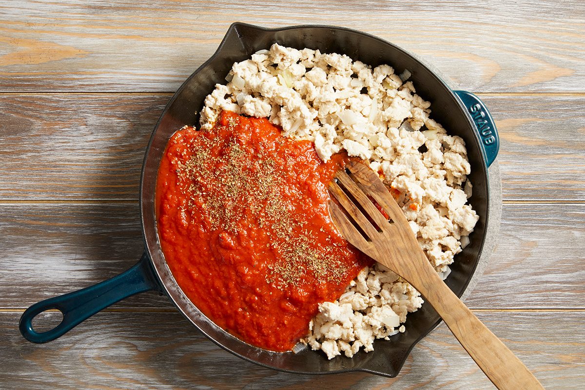 A skillet with cooked ground turkey on one side and tomato sauce with herbs on the other, being stirred together with a wooden spatula on a light wooden surface.