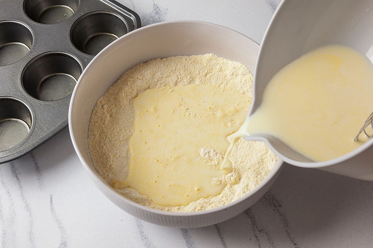 Overhead shot of a bowl of dry cornbread mix being combined with a yellow liquid mixture poured from another bowl, with a muffin tin nearby on a white marble surface;