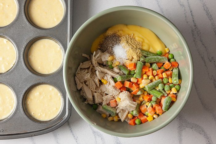 Overhead shot of a bowl with shredded chicken, frozen mixed vegetables, creamy soup, and seasonings beside a muffin tin filled with batter on a marble countertop;