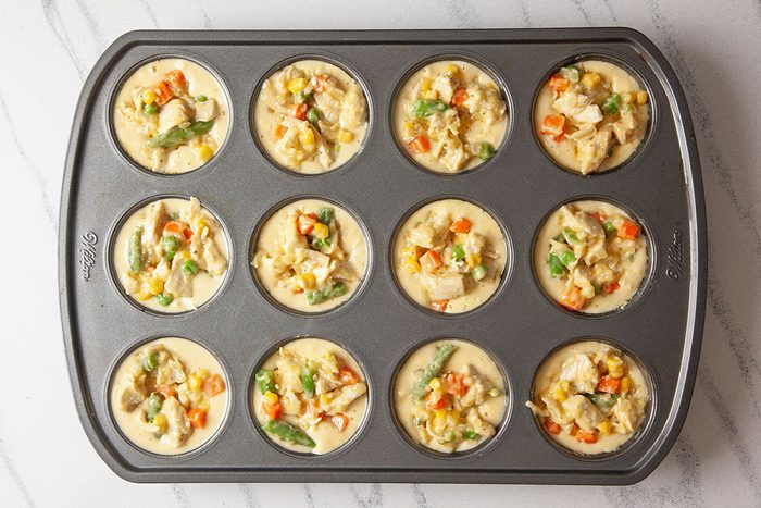 Overhead shot of a muffin tin with 12 cups filled with an uncooked mixture of eggs, vegetables, and chicken, ready to be baked; ingredients include corn, green beans, carrots, and peas;