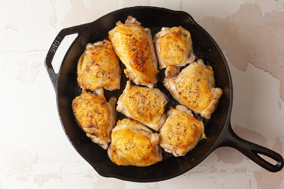 Overhead shot of nine golden-brown, seasoned chicken thighs arranged in a black cast-iron skillet on a light-colored surface