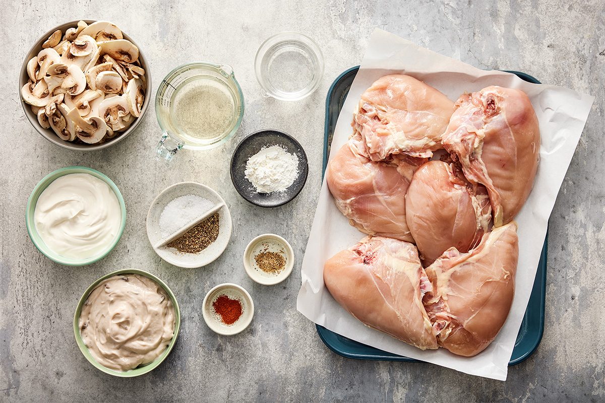 Raw chicken breasts on a tray with bowls of sliced mushrooms, sour cream, flour, spices, oil, and a glass of water arranged on a gray countertop, ready for cooking.