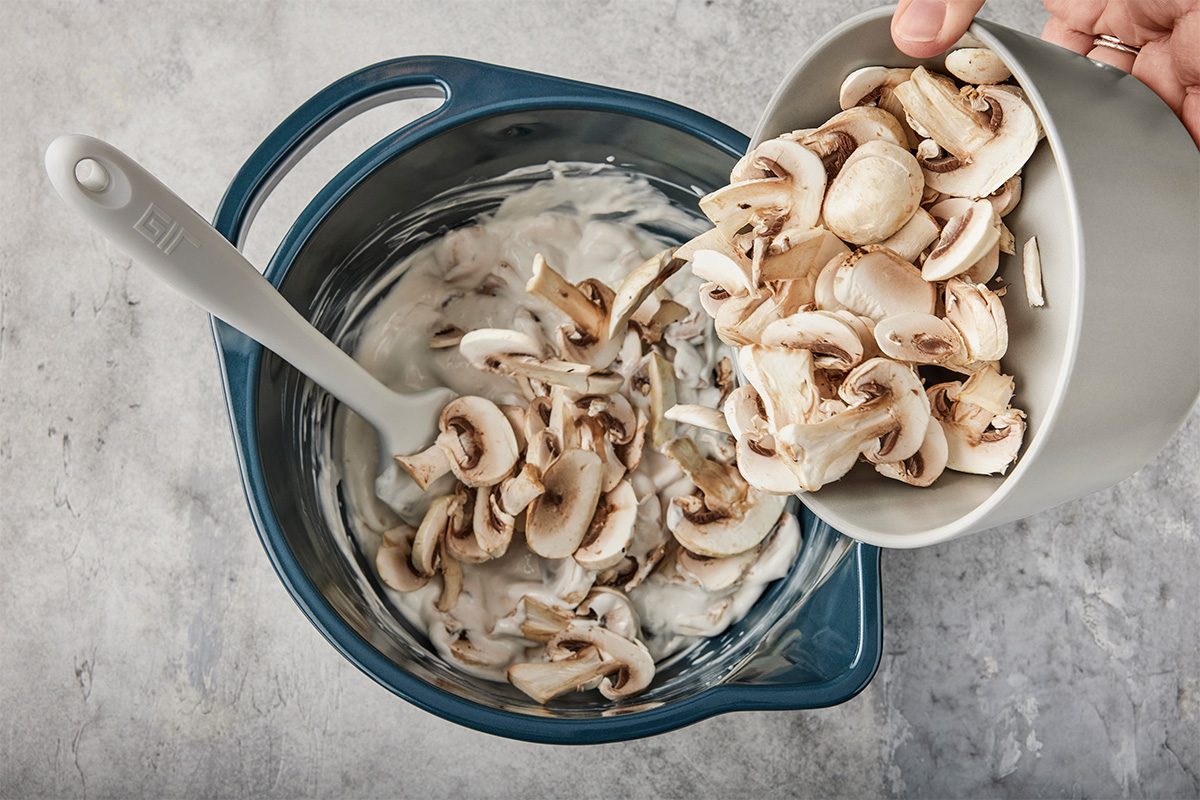 A hand pours sliced mushrooms from a gray bowl into a mixing bowl containing a creamy white mixture, with a spatula resting inside the bowl on a gray countertop.
