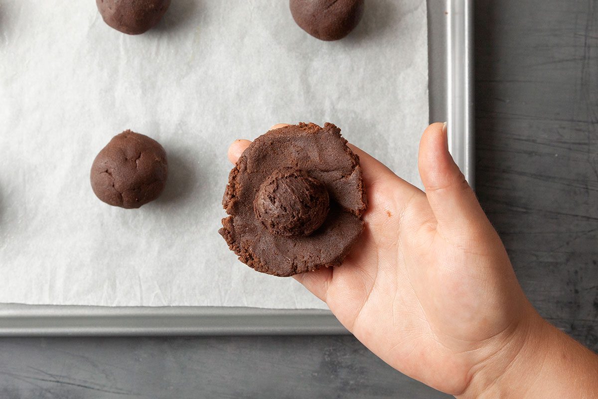 Overhead shot of a hand holds a partially flattened chocolate cookie dough ball above a baking tray lined with parchment paper More round dough balls sit around it The scene shows the cookies being made