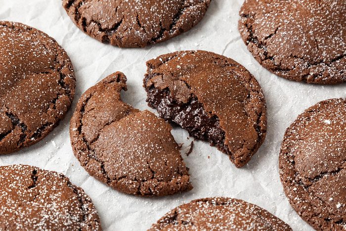 Close-up shot Chocolate Lava Cookies are dusted with powdered sugar and arranged on parchment paper One is broken in half with chocolate oozing out