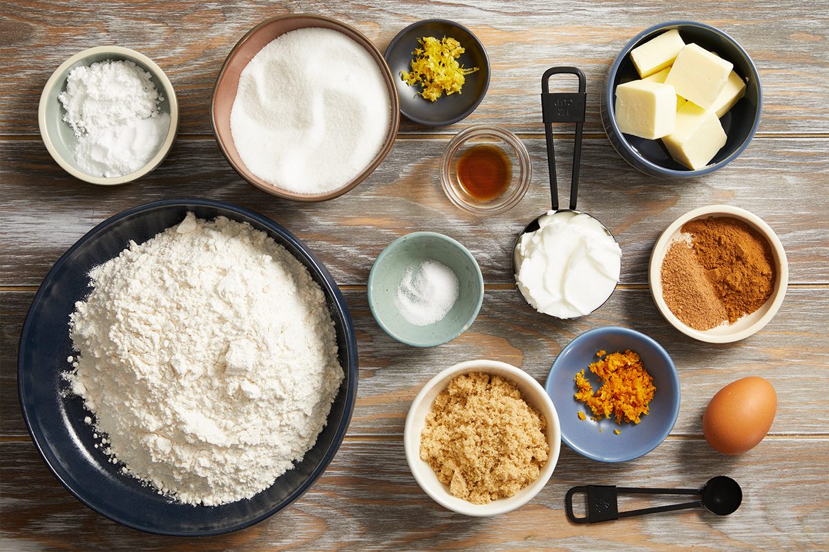 Overhead shot of individual baking ingredients in small bowls and measuring cups, including flour, sugar, brown sugar, butter, eggs, vanilla, and cinnamon, arranged neatly on a wood surface.
