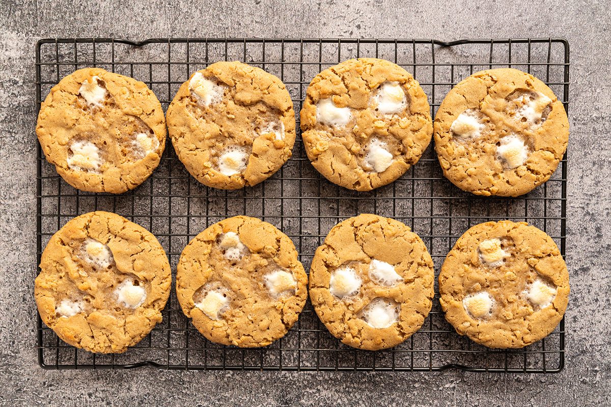 Eight golden brown cookies with white marshmallow pieces are arranged in two rows on a black wire cooling rack, set on a gray textured surface.