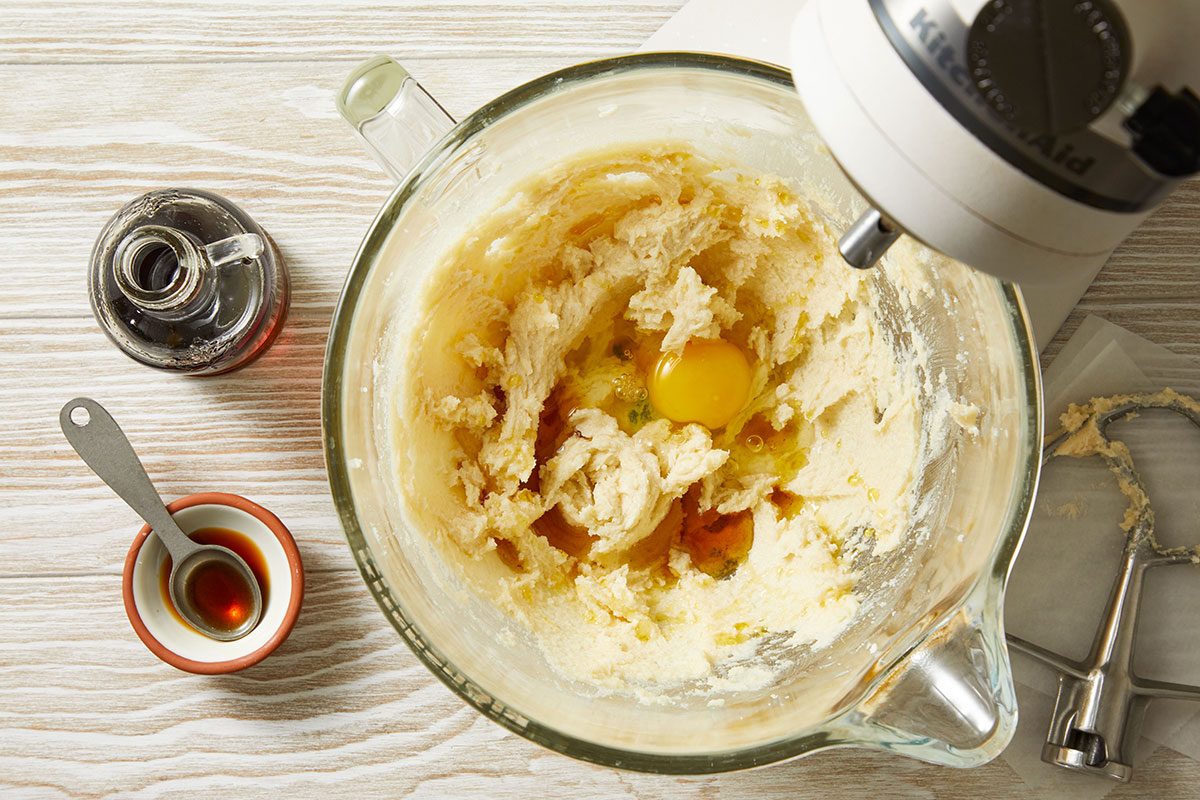 Overhead shot of a stand mixer holds creamed batter with an egg and vanilla in a glass bowl nearby vanilla extract a measuring spoon and a beater rest on a light wooden surface everything is ready for mixing