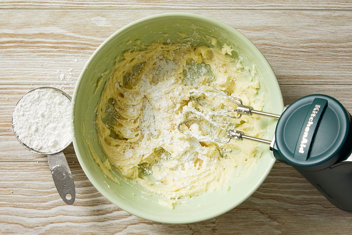 Overhead shot of a mixing bowl has creamy batter and powdered sugar being blended with a hand mixer and a measuring cup filled with powdered sugar is beside it
