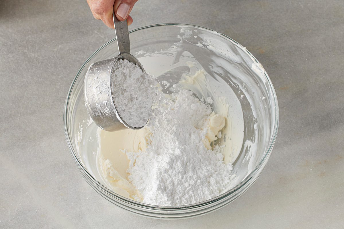 Overhead shot of a hand pouring powdered sugar from a metal measuring cup into a glass bowl with a creamy mixture on a marble surface