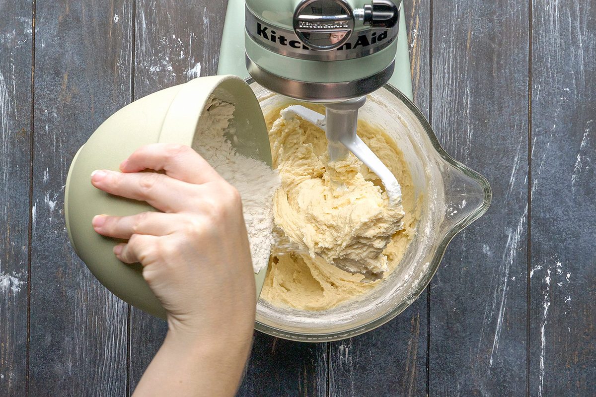 Overhead shot of a person pouring flour from a green bowl into a stand mixer containing creamy batter