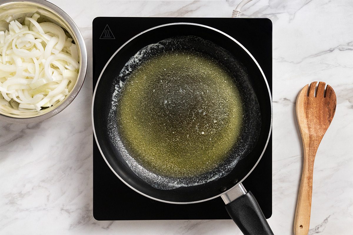 Overhead shot of a large cast-iron or other heavy skillet placed on induction; nearby a wooden spoon and a bowl of onion is visible; all set on a white marble surface;