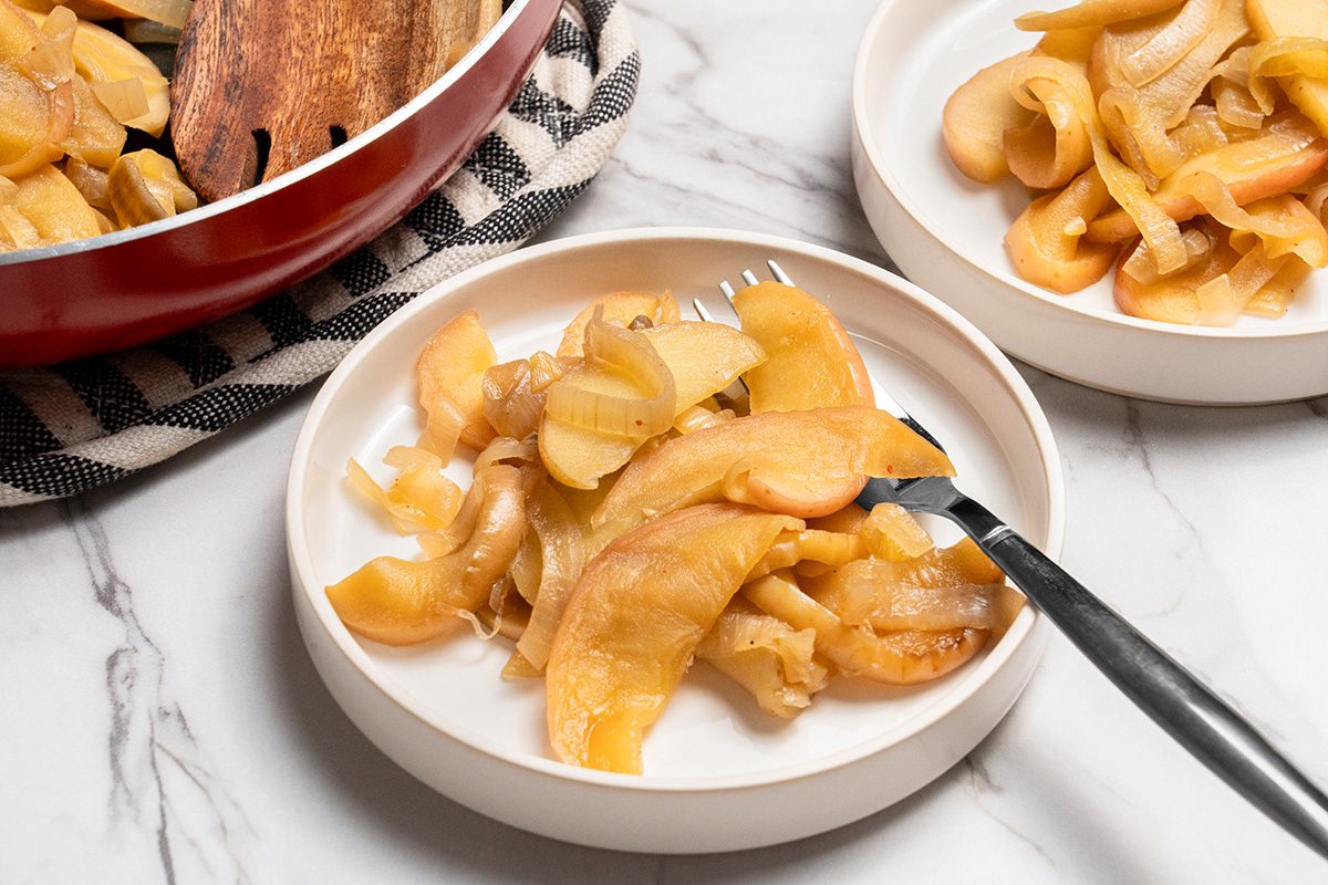 3/4 view angle shot of Fried Onions and Apples served in plates with a fork; a skillet is visible nearby; all set on a white marble surface;