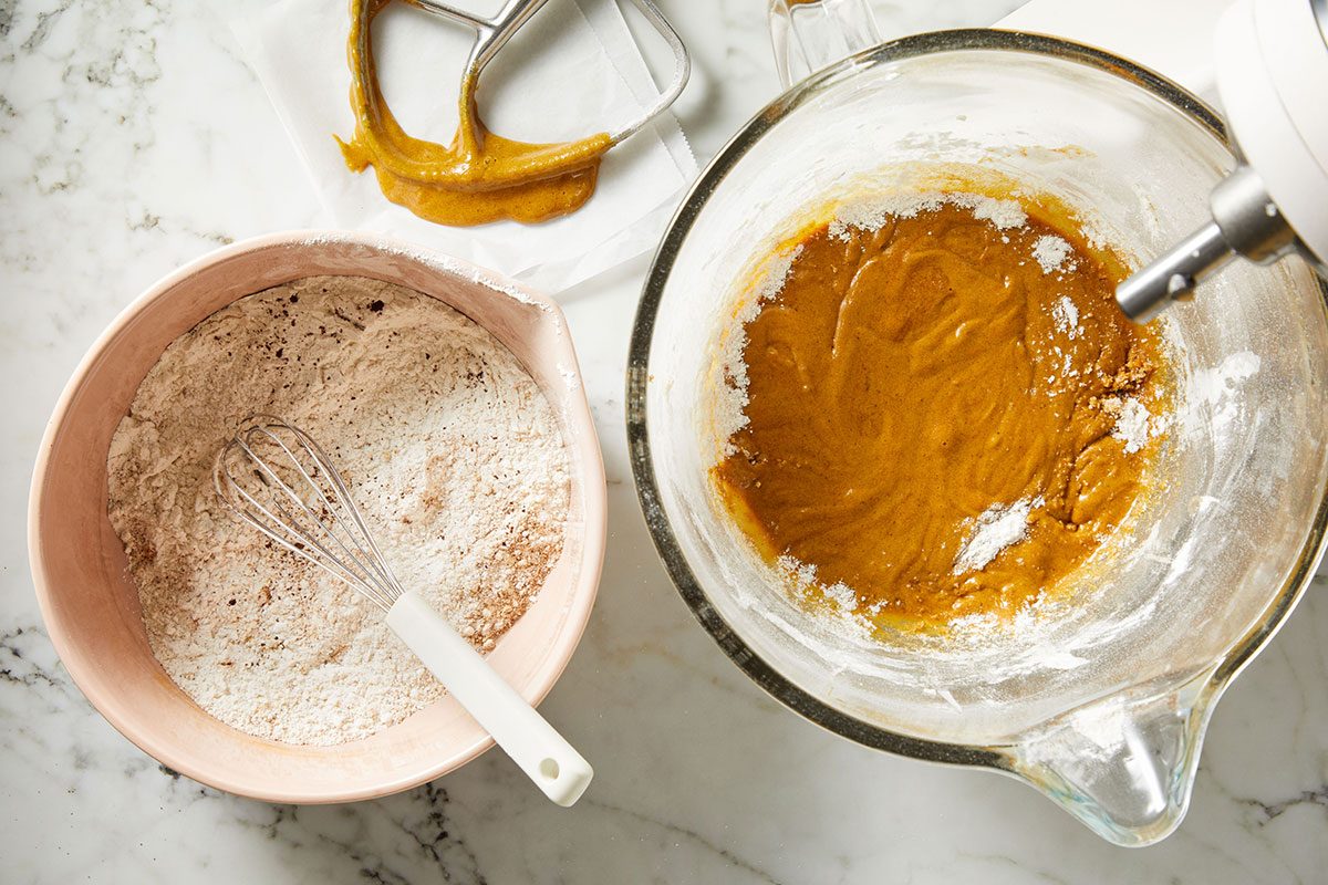 Overhead shot of a glass bowl holds brown batter beside a pink bowl of flour and a whisk on marble A mixer with dough on the paddle is nearby