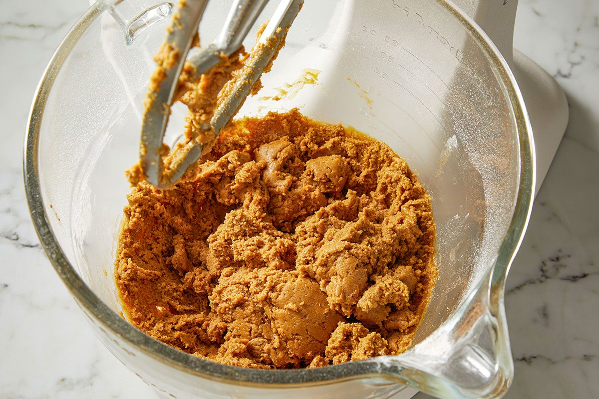 Overhead shot of a glass bowl holds brown batter beside a pink bowl of flour and a whisk on marble A mixer with dough on the paddle is nearby