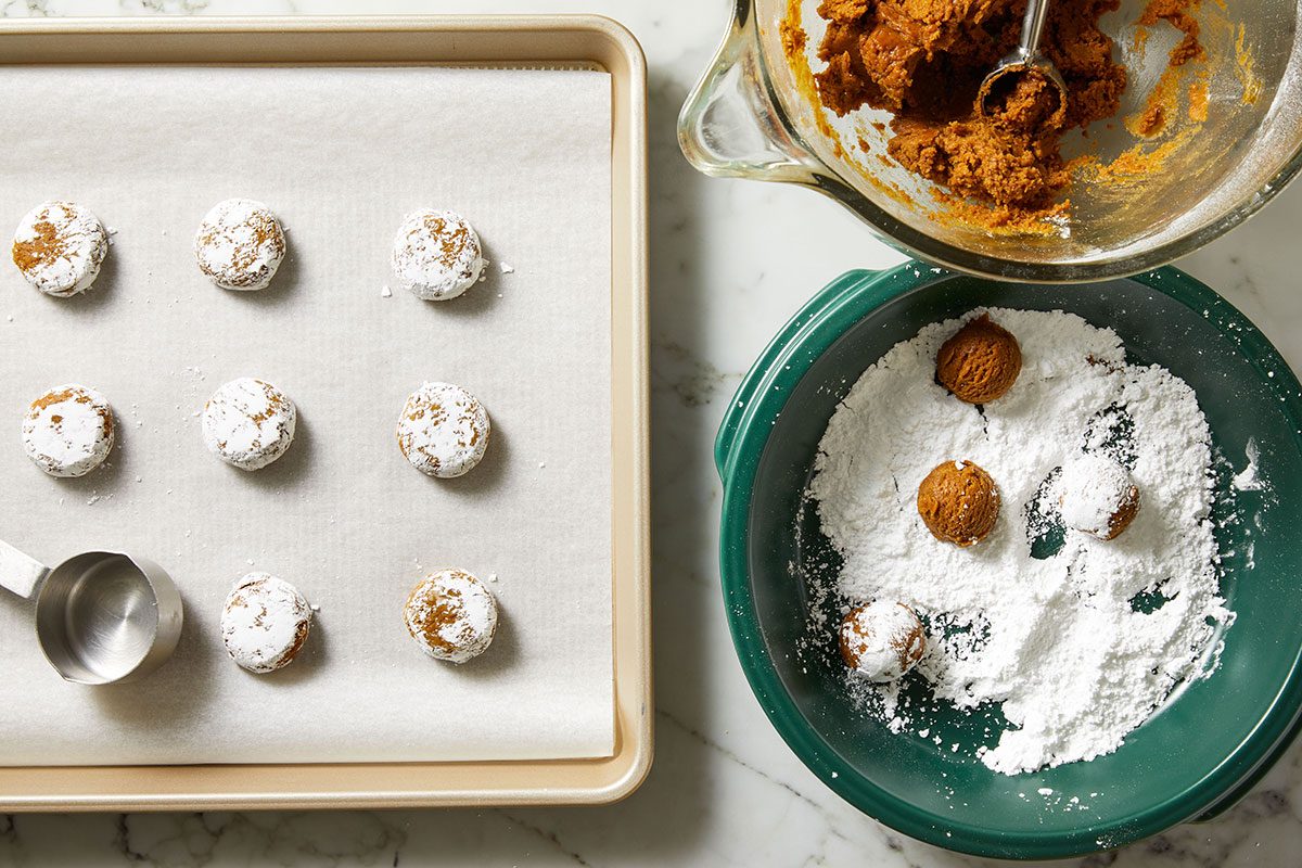 Overhead shot of a baking tray holds dough balls dusted with powdered sugar and it sits next to a bowl of dough and one of powdered sugar on marble with some dough balls being coated in sugar