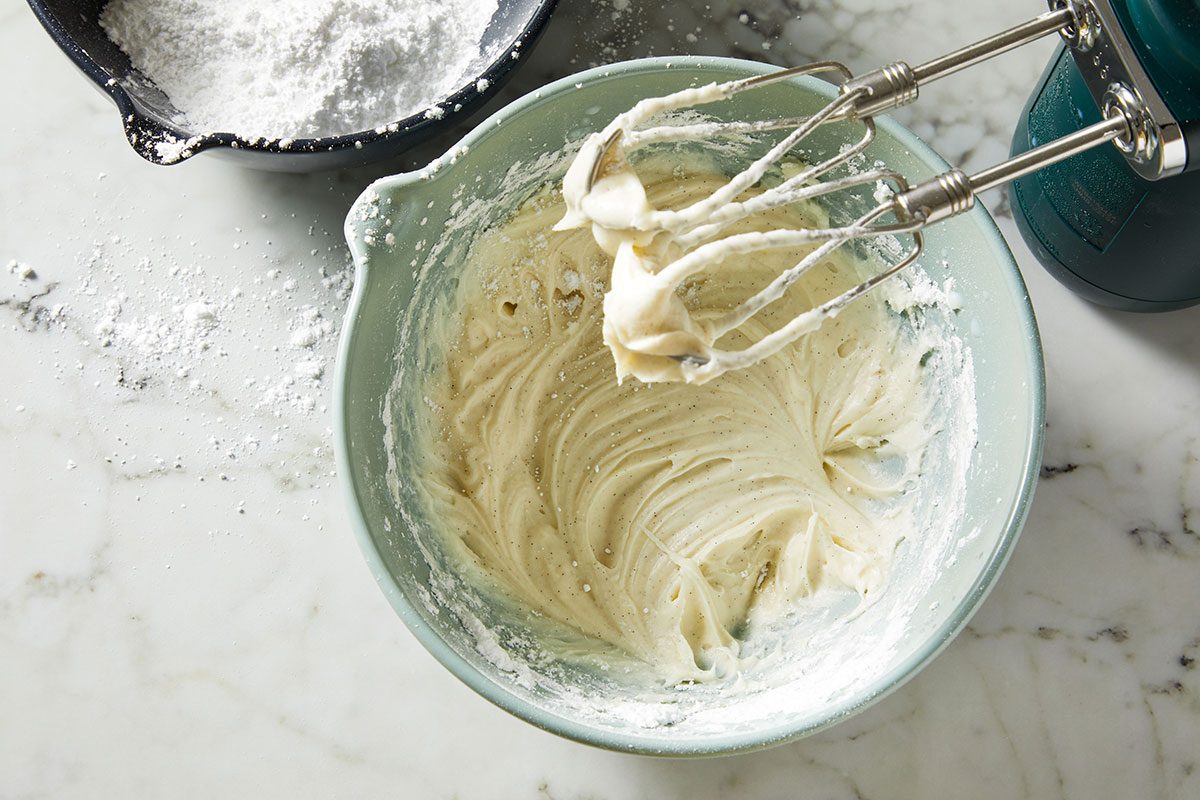 Overhead shot of a mixing bowl holds creamy batter as electric beaters work on it and it sits on a marble countertop with powdered sugar close by
