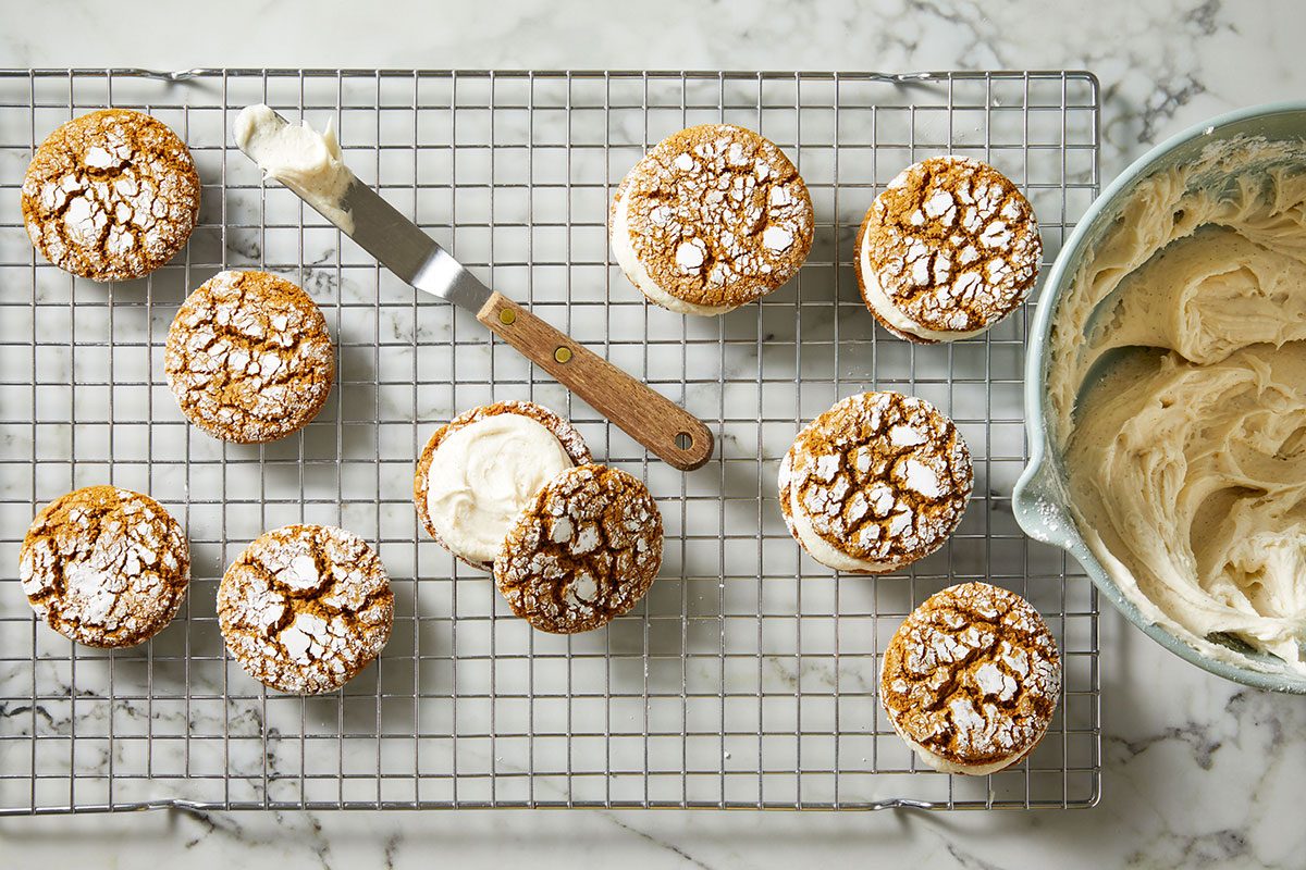 Overhead shot of Cookies dusted with powdered sugar rest on a cooling rack and some are sandwiched with creamy filling a spatula and a bowl of frosting sit on the right