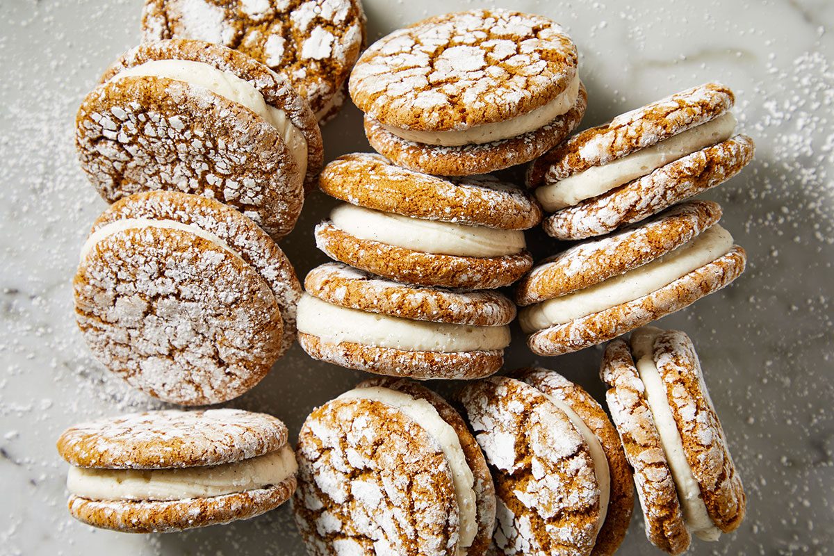 Close-up shot of Gingerbread Crinkle Cookie Sandwiches are displayed on a light marble surface with some cookies stacked and others laying flat and the tops are crackled and dusted with powdered sugar and creamy white filling shows between the cookies