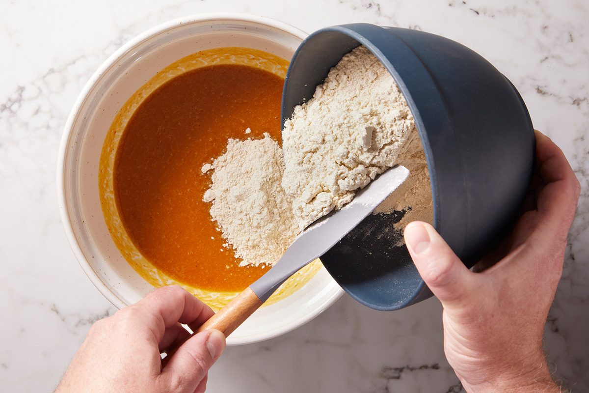 A person pours flour from a blue bowl into a large white bowl containing a yellow-orange liquid mixture, using a spatula to help transfer the flour. Both bowls are on a white countertop.