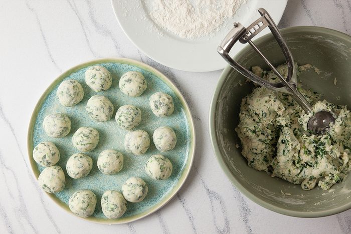 Overhead shot of a plate of uncooked spinach and ricotta dumplings beside a bowl of dough and a scoop on a white countertop, with a plate of flour in the background