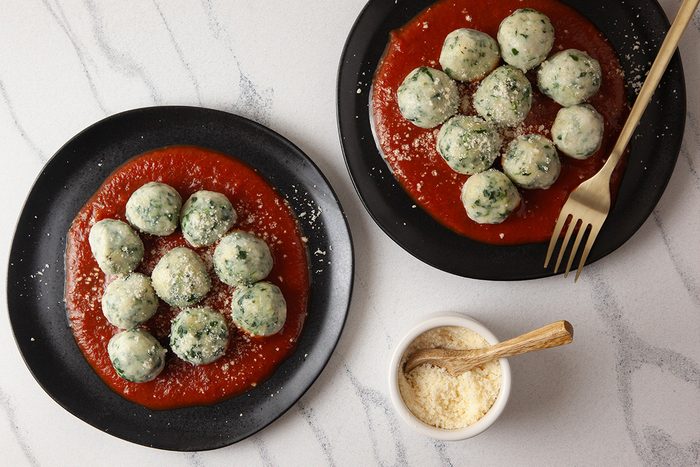 Overhead shot of two black plates of gnudi on tomato sauce topped with grated cheese, with a gold fork on one plate and a small bowl of cheese nearby on a marble surface