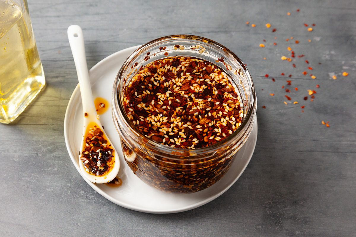 3/4 shot of a jar of Chili Crisp with a spoon resting inside, showing the chunky mixture of fried garlic, chili flakes, and oil.