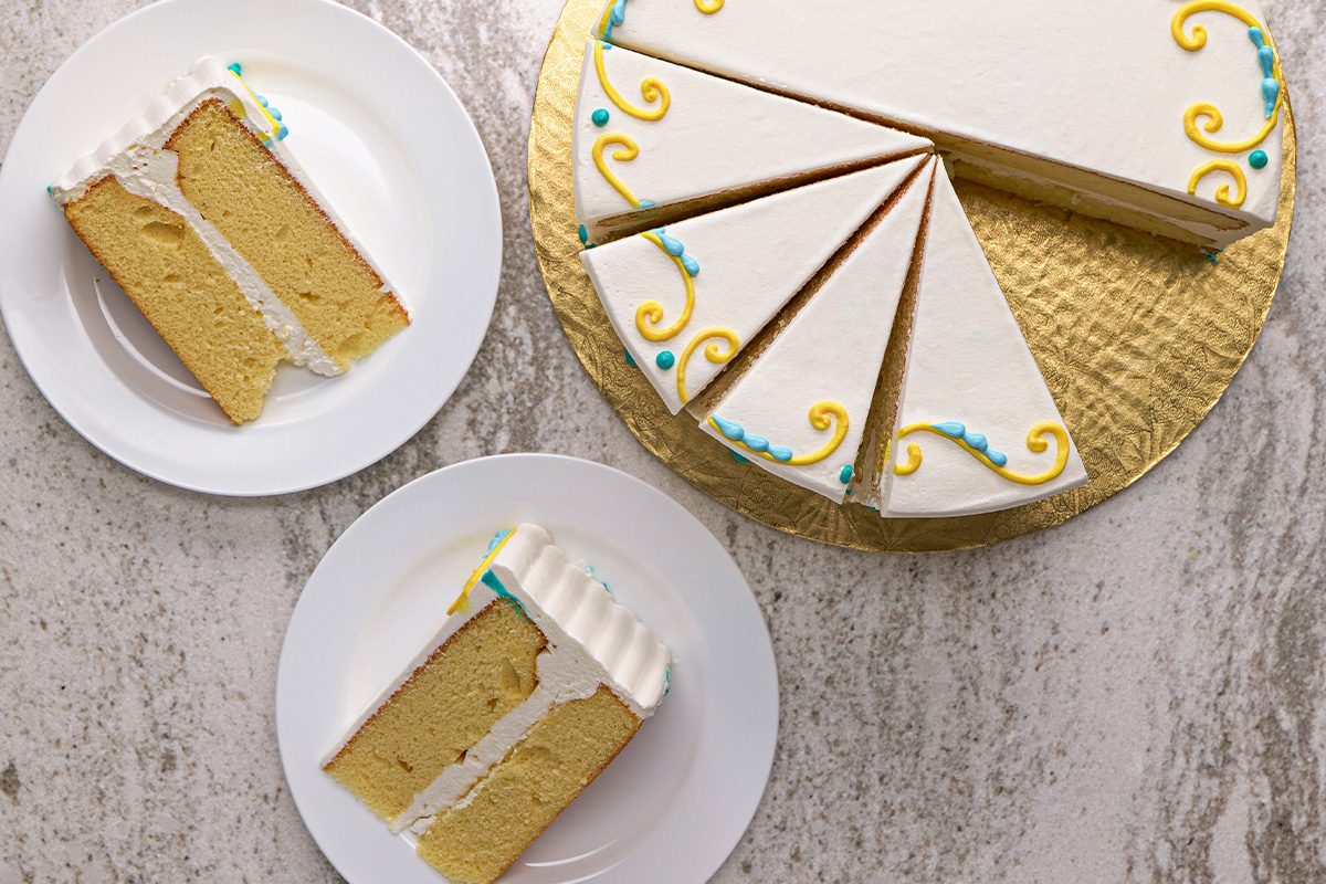 A round yellow cake with white frosting and colorful decorative swirls is sliced; two individual cake slices are served on white plates next to the remaining cake on a gold tray.