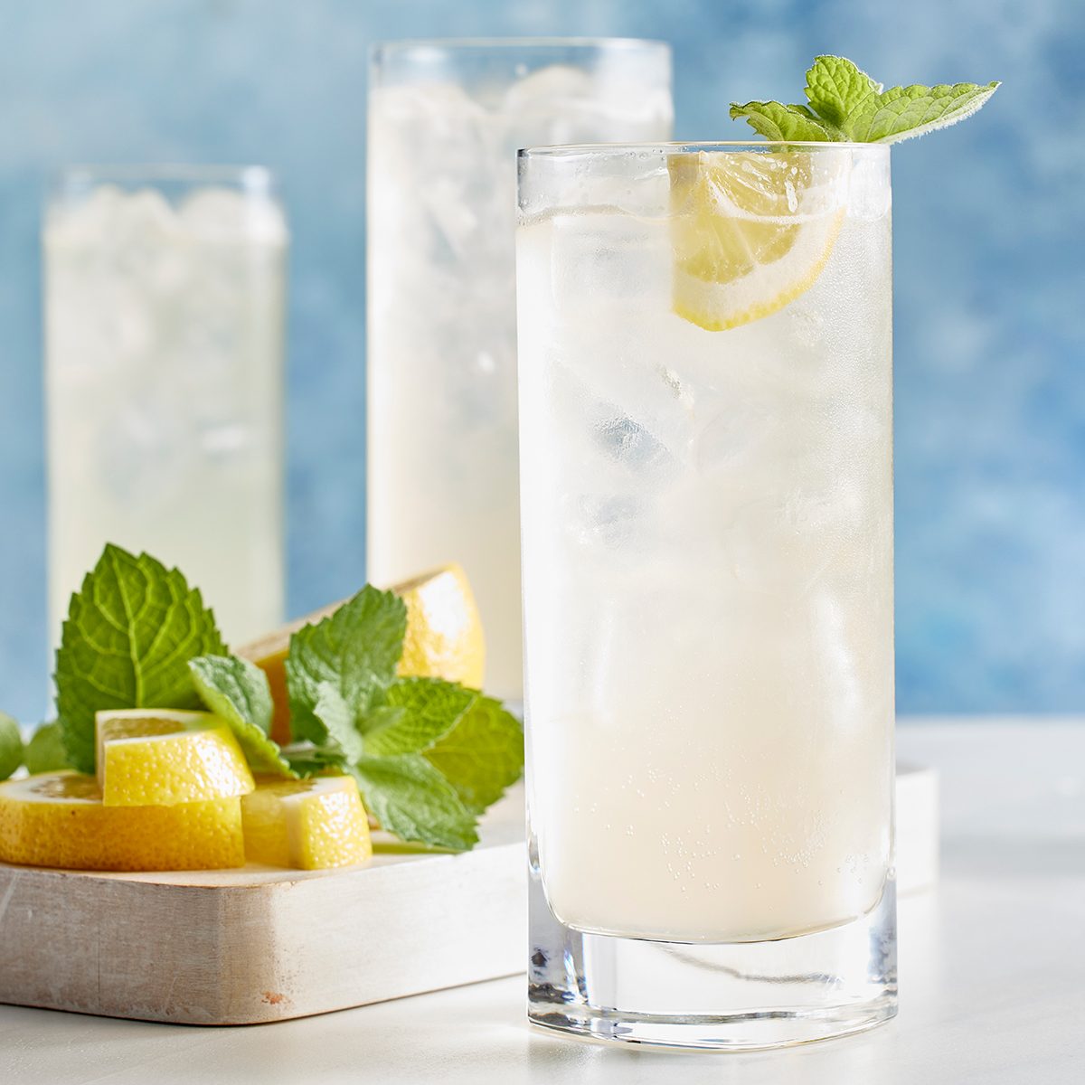 A tall glass of lemonade with ice, garnished with a lemon slice and mint, sits on a table beside a wooden tray holding lemon wedges and fresh mint leaves. Two more lemonade glasses are in the background.