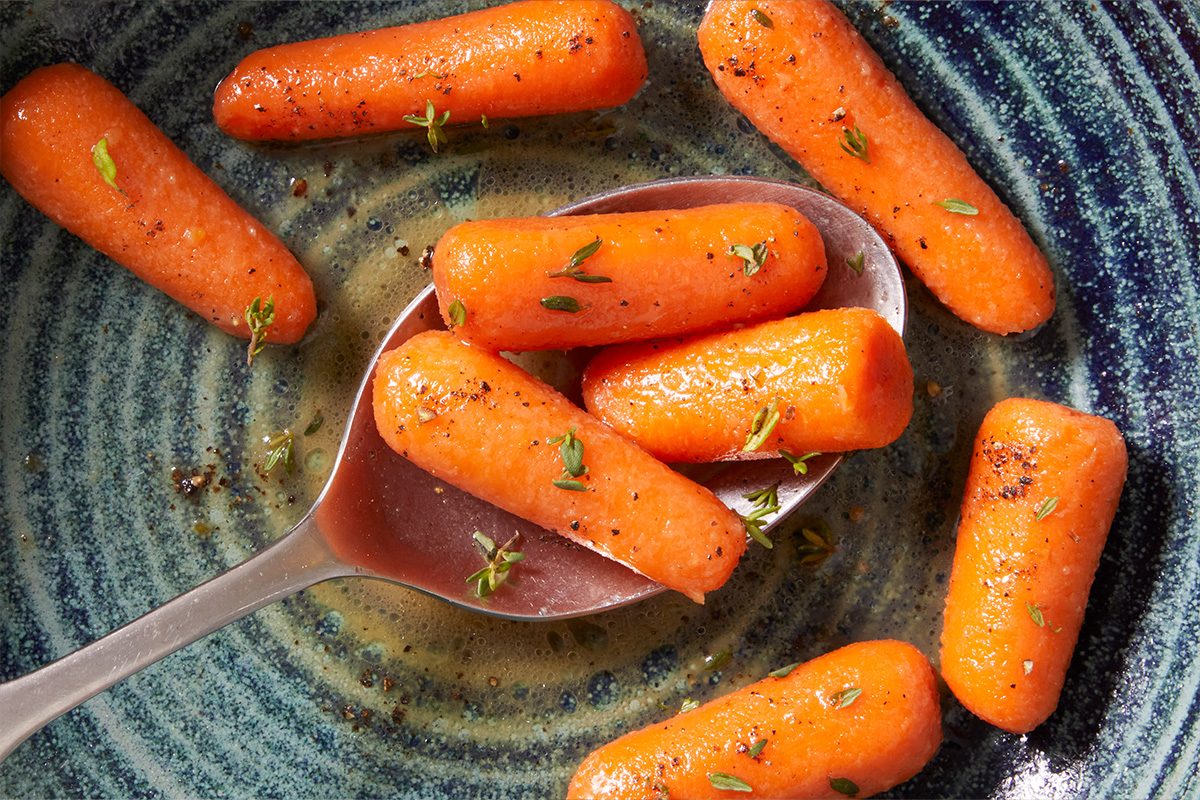 A silver spoon rests on a blue plate filled with bright orange glazed baby carrots, garnished with small green herb pieces and a light sauce.