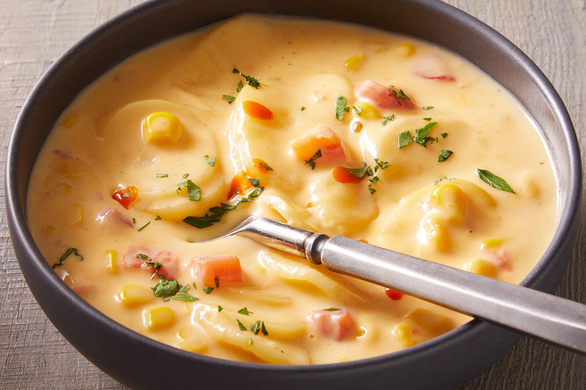 A bowl of creamy soup with visible chunks of potatoes, corn, carrots, and herbs, served with a spoon in the bowl.